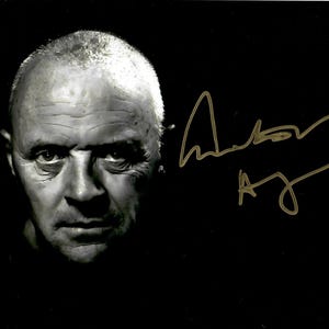 May include: Black and white portrait of a man with a serious expression. The image is signed in gold ink. The man's face is in sharp focus, with the background fading to black. The signature is in cursive.