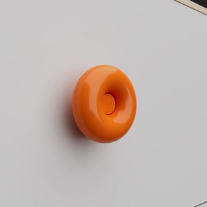 Peut inclure: Un bouton de meuble circulaire orange vif avec une finition lisse et brillante et un bouton central encastré. Le bouton est monté sur une surface blanche plane, présentant une forme simple et arrondie rappelant un beignet.