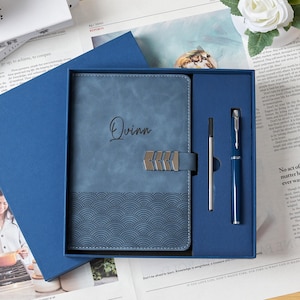 May include: A blue gift set featuring a personalized journal with the name "Quinn" and a matching pen. The journal has a wave pattern at the bottom and a silver clasp. The set is presented in a blue box.