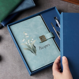 May include: A light blue journal with a floral design and the name "Harper" is displayed in a blue gift box. The journal has a decorative clasp and comes with a pen and a pencil. Other journals are visible in the background.