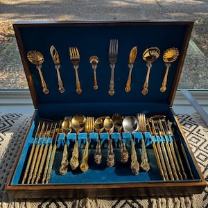 Puede incluir: Un juego de cubiertos vintage en tono dorado exhibido en un estuche de madera con interior de terciopelo azul. El juego incluye cucharas, tenedores y cuchillos, dispuestos ordenadamente. El estuche está abierto, revelando el servicio completo.