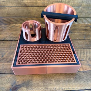 May include: A copper-colored espresso station with a knock box, portafilter holder, and a drip tray with a honeycomb pattern. The knock box has a black bar. The portafilter holder has oval cutouts. The drip tray is 10 inches wide.
