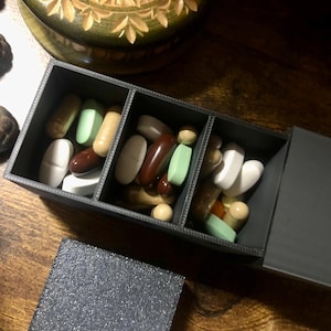 May include: A dark gray pill organizer with three compartments, filled with various pills and capsules in white, brown, green, and beige. The lid is open, revealing the contents. The organizer sits on a wooden surface.