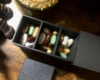 May include: A dark grey pill organiser with three compartments, each filled with various colourful pills and capsules. The lid is partially open, revealing the contents. The pills are in shades of white, brown, green, and beige.