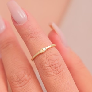 Könnte beinhalten: Ein goldfarbener Ring mit einem kleinen augenförmigen Design. Der Ring wird an einem Finger getragen, vor einem weichen, neutralen Hintergrund. Das Design ist minimalistisch und zentriert.