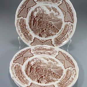 May include: Two white ceramic plates with a brown transferware design. The plates feature a nautical scene with a ship and figures, surrounded by decorative borders and text. The plates are displayed on a clear stand.