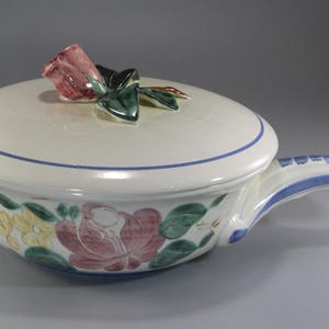 May include: Cream-colored ceramic casserole dish with a lid, featuring a floral design in pink, yellow, and green. The lid is topped with a rose and green leaves. The handle is white with blue accents.