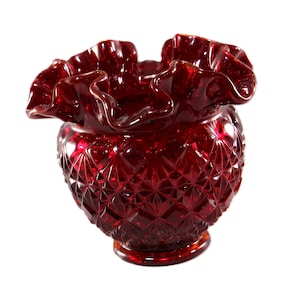 May include: A ruby red glass vase with a ruffled rim and diamond-cut pattern. The vase has a round base and a bulbous body. The ruffled rim adds a decorative touch to the piece. The glass is a deep, rich red color.