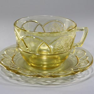 May include: A vintage yellow glass teacup and saucer set. The cup and saucer have a decorative embossed floral and geometric design. The cup has a handle and sits on a matching saucer. The set is made of transparent yellow glass.