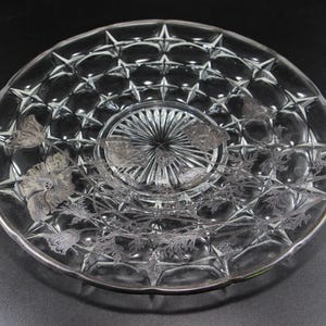 May include: A clear glass plate with a starburst pattern in the center and a floral design around the edges. The plate has a silver rim and is likely used for serving or display.