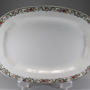 May include: Oval white serving platter with a floral border. The border features pink, yellow, and red flowers with green leaves on a black and gold background. The platter is supported by a clear stand.