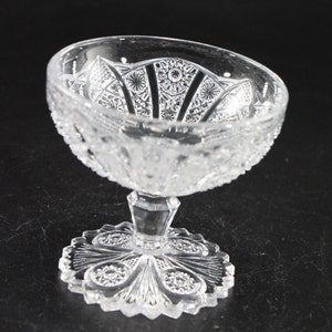 May include: A clear, cut-glass dessert dish with a detailed, ornate pattern. The bowl has a scalloped edge and sits atop a pedestal base with a decorative, circular foot. The glass is transparent, allowing light to pass through the intricate designs.