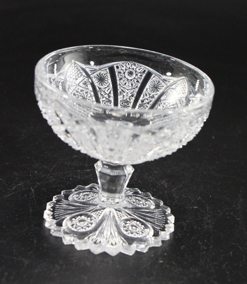 May include: A clear, cut-glass dessert dish with a detailed, ornate pattern. The bowl has a scalloped edge and sits atop a pedestal base with a decorative, circular foot. The glass is transparent, allowing light to pass through the intricate designs.