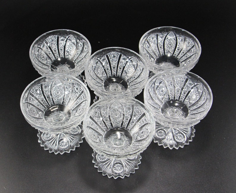 May include: Six clear glass dessert bowls with intricate cut crystal designs. Each bowl has a rounded shape, a pedestal base, and detailed patterns. The bowls are arranged on a black surface, showcasing their elegant craftsmanship.