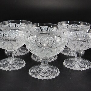 May include: Six clear, cut-glass dessert bowls with ornate, starburst-like patterns. Each bowl sits atop a short stem and a decorative, scalloped base. The bowls are arranged on a dark surface, showcasing their intricate details.