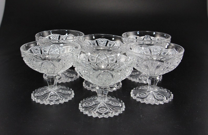 May include: Six clear, cut-glass dessert bowls with ornate, starburst-like patterns. Each bowl sits atop a short stem and a decorative, scalloped base. The bowls are arranged on a dark surface, showcasing their intricate details.