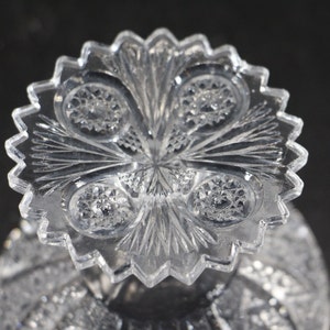 May include: Close-up of a clear, cut-glass object with a starburst pattern. The object has a scalloped edge and intricate detailing, including circular and linear designs. The glass reflects light, creating a sparkling effect. The object is likely a decorative piece or part of a larger item.