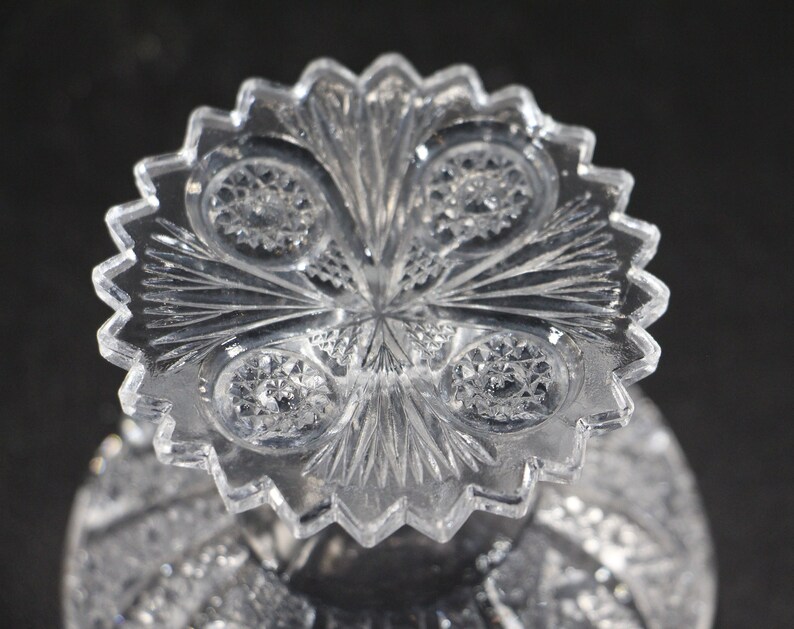 May include: Close-up of a clear, cut-glass object with a starburst pattern. The object has a scalloped edge and intricate detailing, including circular and linear designs. The glass reflects light, creating a sparkling effect. The object is likely a decorative piece or part of a larger item.