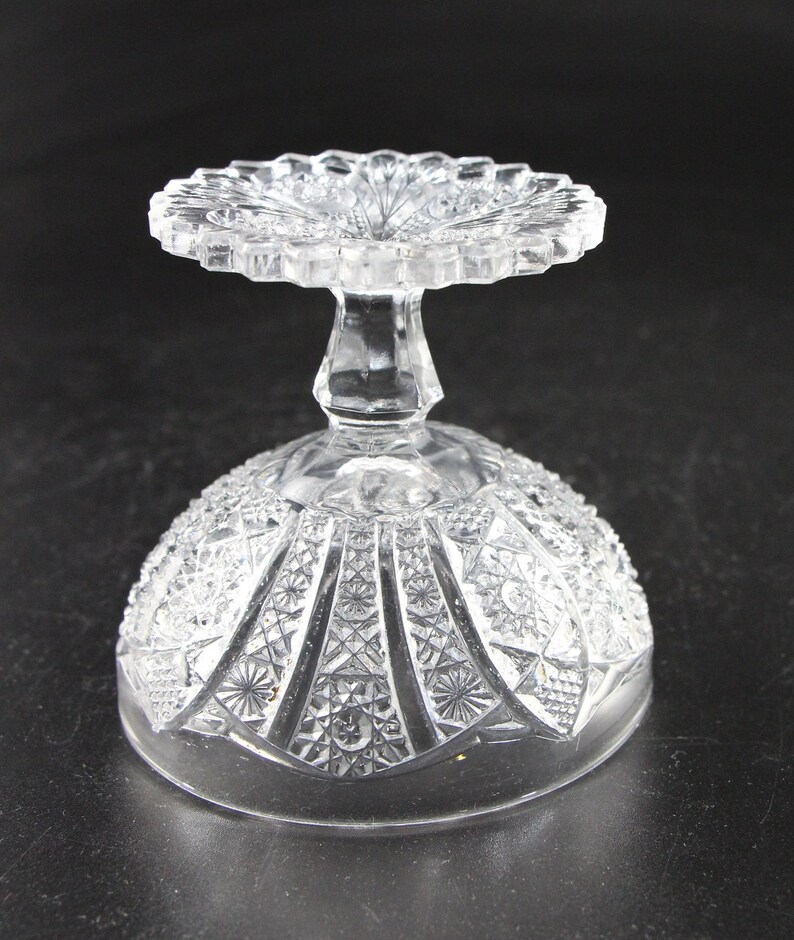 May include: A clear, vintage-style glass dessert dish with a detailed, textured pattern. The dish features a round, scalloped top and a pedestal base. The bowl has a geometric design, and the glass is transparent, allowing light to pass through.