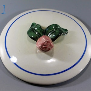 May include: A round, off-white ceramic lid with a blue border and a decorative rose and leaf handle. The rose is pink and the leaves are green. The text "# 1" is in blue in the upper left corner.