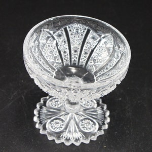 May include: A clear, ornate glass dessert dish with a detailed, cut-glass pattern. The bowl sits atop a pedestal base with a decorative, starburst-like design. The glass has a textured, sparkling appearance, reflecting light.