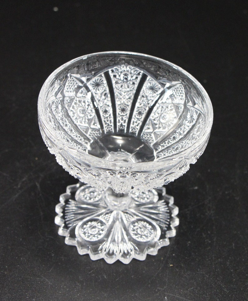 May include: A clear, ornate glass dessert dish with a detailed, cut-glass pattern. The bowl sits atop a pedestal base with a decorative, starburst-like design. The glass has a textured, sparkling appearance, reflecting light.