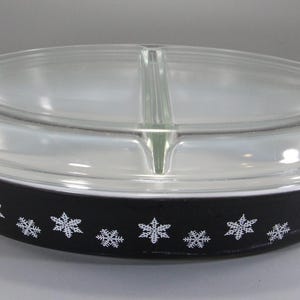 May include: Black oval casserole dish with a clear glass lid. The dish has white snowflake designs around the rim. The lid is divided in the middle. The dish has handles on each side.