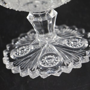 May include: Close-up of a clear, cut-glass pedestal dish. The base has a scalloped edge with a detailed, fan-like pattern. The stem is faceted, and the dish's rim is also textured. The glass reflects light, creating a sparkling effect.