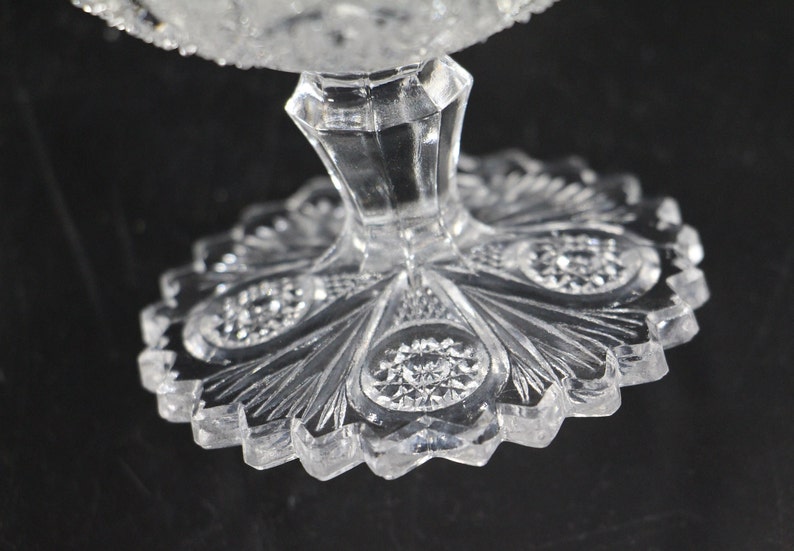 May include: Close-up of a clear, cut-glass pedestal dish. The base has a scalloped edge with a detailed, fan-like pattern. The stem is faceted, and the dish's rim is also textured. The glass reflects light, creating a sparkling effect.