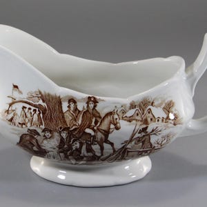May include: White ceramic gravy boat with a brown transferware design depicting a historical winter scene with figures, houses, and a flag. The gravy boat has a spout, a handle, and a pedestal base.