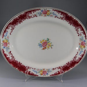 May include: A white oval serving platter with a red and gold border. The border is decorated with a floral pattern. The center of the platter is plain white.