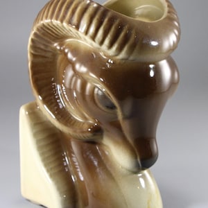 May include: A ceramic ram head vase in shades of brown and beige. The vase features a detailed depiction of a ram's head with curved horns and a textured surface. The vase has a glossy finish and a hollow interior.