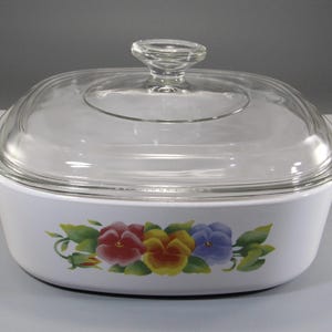 May include: White casserole dish with a clear glass lid and handle. The dish features a floral design of red, yellow, and purple pansies with green leaves. The lid has a clear glass knob.