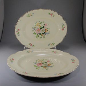 May include: Two oval ceramic serving platters with a floral design. The platters are cream colored with a gold trim and feature a bouquet of pink, yellow, and blue flowers.