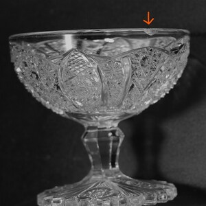 May include: Clear, cut-glass dessert bowl with a detailed, symmetrical pattern. The bowl has a wide, decorative rim and sits on a pedestal base. The glass has a slight chip on the rim. The bowl is designed for serving desserts.