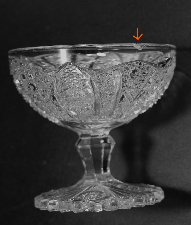 May include: Clear, cut-glass dessert bowl with a detailed, symmetrical pattern. The bowl has a wide, decorative rim and sits on a pedestal base. The glass has a slight chip on the rim. The bowl is designed for serving desserts.