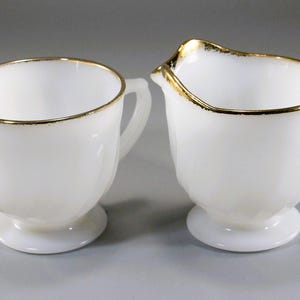 May include: A white milk pitcher and creamer set with gold trim. The creamer has a unique spout design. Both pieces have a pedestal base and a handle. The gold trim adds a touch of elegance to the set.