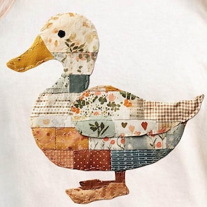 May include: Cream-colored t-shirt featuring a patchwork duck design. The duck has a yellow beak and feet, with a body composed of various fabric patterns including floral, plaid, and solid colors. The shirt is a casual style.