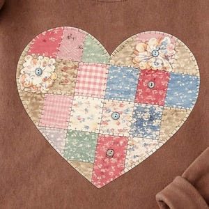 Può includere: Una felpa marrone con un design patchwork a forma di cuore. Il cuore è composto da quadrati di tessuto nei toni del rosa, blu e beige, con motivi floreali e a quadri. La felpa ha un girocollo e maniche lunghe con polsini a coste.