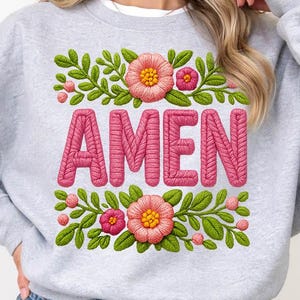 May include: A pale grey sweatshirt featuring the word "AMEN" in pink, embroidered lettering. The word is framed by a floral design with pink flowers and green leaves. The sweatshirt is a casual style.