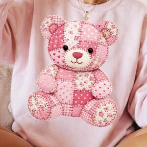 May include: A light pink sweatshirt featuring a patchwork teddy bear design. The bear is composed of various pink fabrics with floral and polka dot patterns. The sweatshirt has a crew neck and long sleeves.