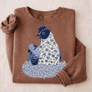 Può includere: Una felpa marrone con un disegno di gallina floreale blu e bianco. La gallina è seduta su un nido con lo stesso motivo floreale. La felpa ha maniche lunghe e girocollo.