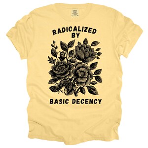 Op de afbeelding: Een lichtgeel t-shirt met de zwarte tekst "RADICALIZED BY BASIC DECENCY". Het shirt heeft een zwart bloemendesign met gedetailleerde bloemen en bladeren.
