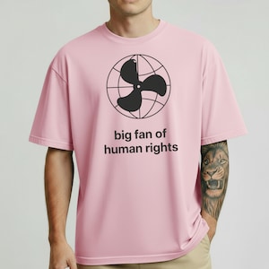Op de afbeelding: Lichtroze t-shirt met een zwarte afbeelding van een ventilator in een wereldbol en de tekst "big fan of human rights". Het shirt heeft korte mouwen en een ronde hals.