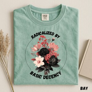 May include: A pale green t-shirt featuring a floral design within a circle. The design incorporates pink, red, and black flowers. The text "RADICALIZED BY BASIC DECENCY" is curved above and below the floral arrangement.