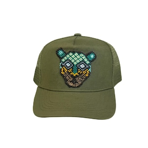 May include: An olive green trucker hat with a beaded owl design. The owl is made with teal, yellow, and brown beads. The hat has a mesh back and a curved peak.