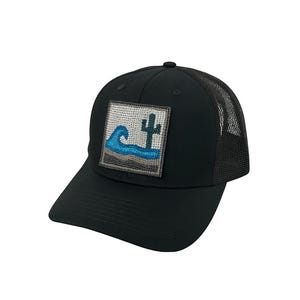 Puede incluir: Gorra de béisbol negra con parte trasera de malla y visera curva. La gorra presenta un parche cuadrado con un diseño de paisaje desértico, que incluye una ola azul y un cactus. El diseño está bordado en tonos azules, grises y blancos.