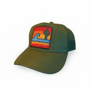 Puede incluir: Gorra de camionero verde oliva con parte trasera de malla y visera curva. La parte delantera presenta un parche cuadrado con un diseño de paisaje desértico, que incluye un cactus, olas y una puesta de sol en tonos rojos, naranjas y azules.