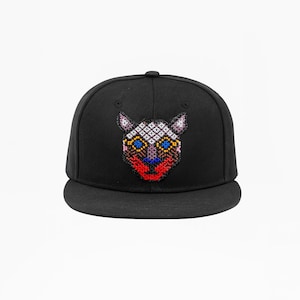 Könnte beinhalten: Schwarze Baseballkappe mit einem farbenfrohen, perlenbesetzten Design eines stilisierten Katzenkopfes. Der Katzenkopf hat blaue Augen, eine rote Nase und ein weißes und rosa gemustertes Gesicht. Die Kappe hat einen flachen Schirm.