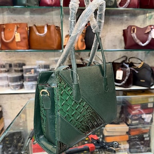 Puede incluir: Un bolso de mano verde oscuro con un acabado texturizado y un acento con estampado de cocodrilo. El bolso tiene forma rectangular con un asa superior y una correa para el hombro desmontable. Otros bolsos de mano de varios colores son visibles en estantes en el fondo.